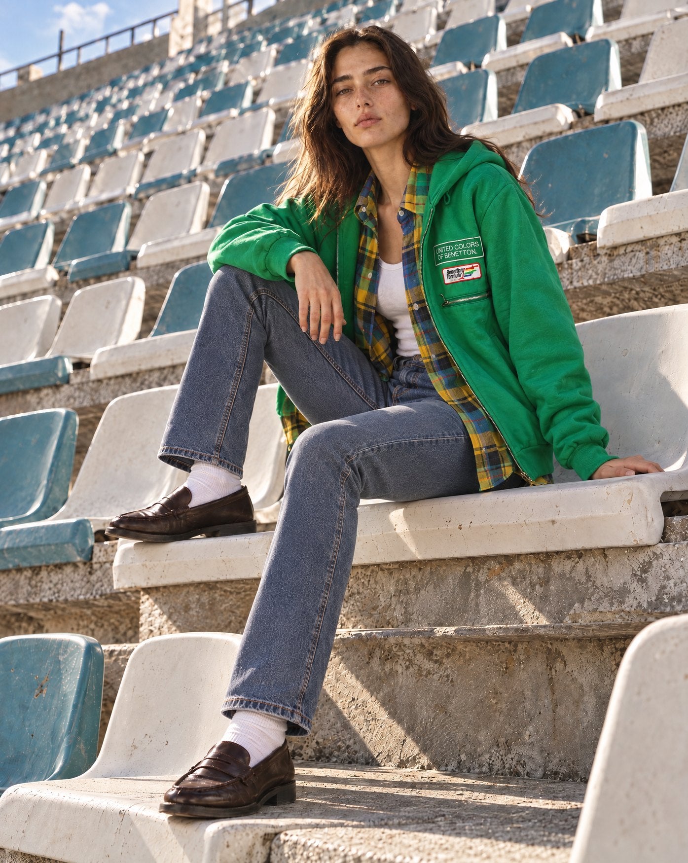 Woman on stadium bleachers in a green Benetton zip hoodie over a yellow plaid flannel shirt, jeans and loafers
