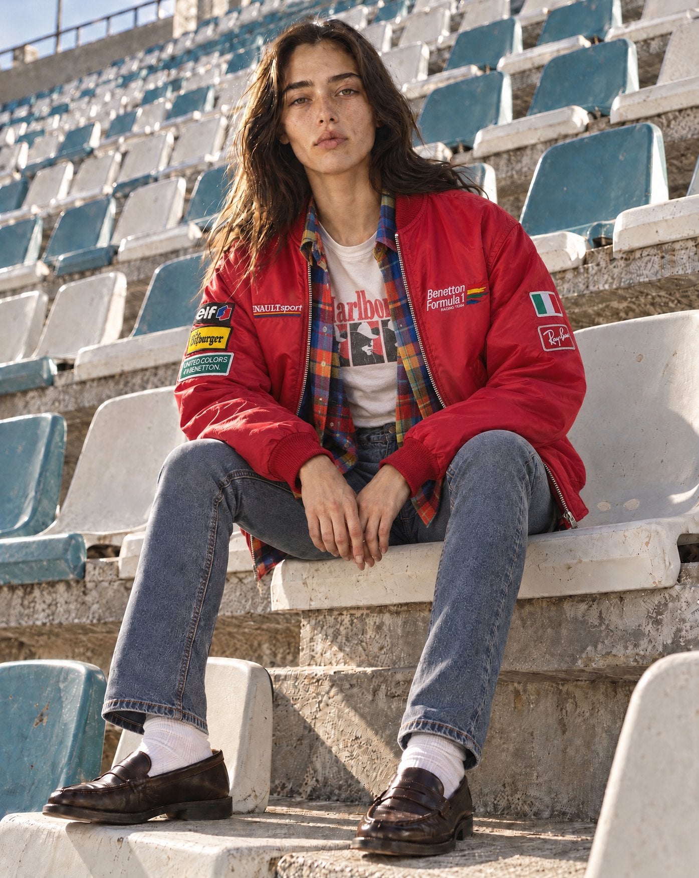 Woman seated on stadium bleachers in a red Benetton Formula 1 bomber over a plaid flannel shirt, jeans and loafers